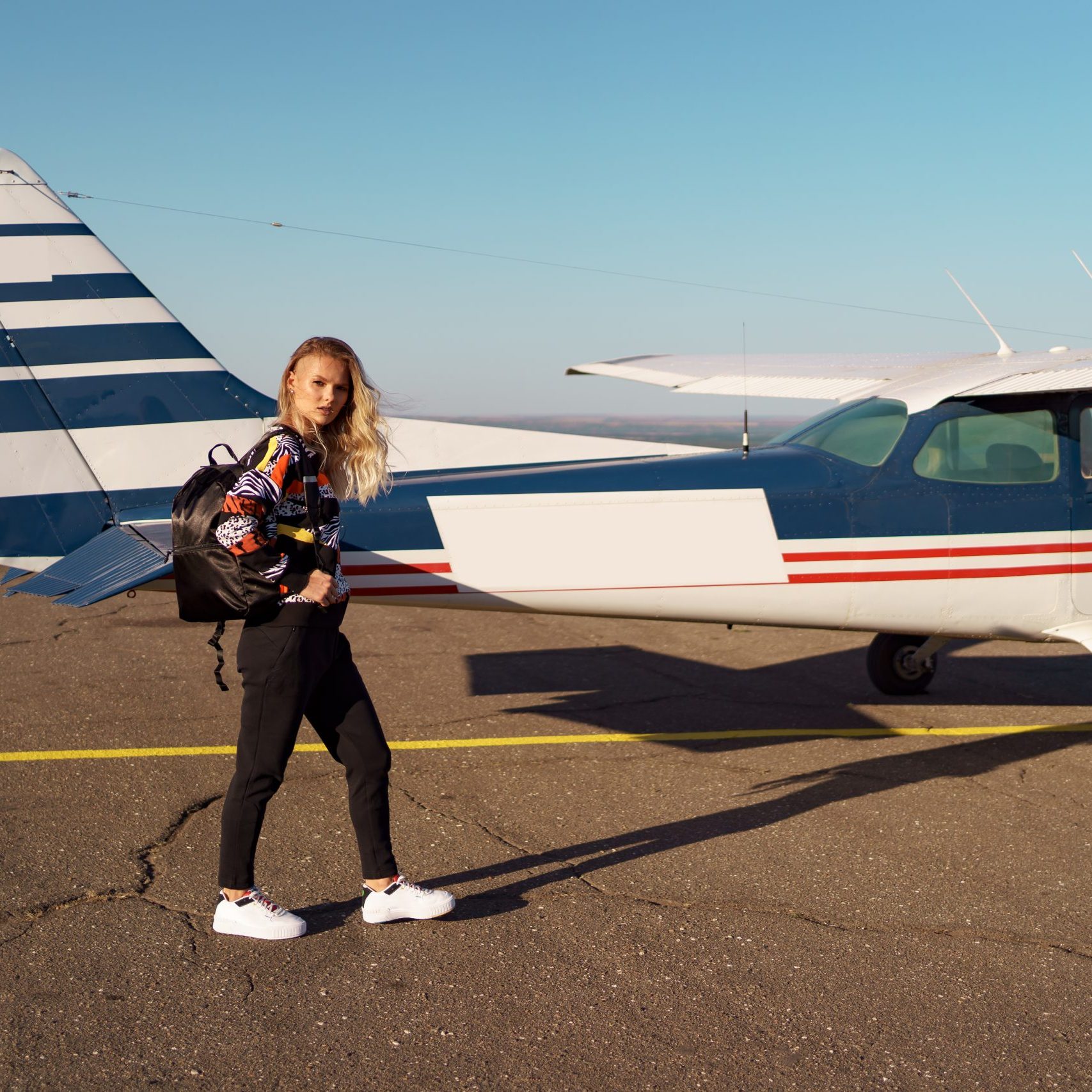Young woman model with a modern haircut posing near a private plane wearing trendy casual outfit and black backpack
